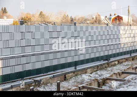 Stacked concrete blocks behind a metal fence with snow on the ground, exposed trenches, wooden supports, and houses with sloped roofs in the backgroun Stock Photo