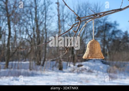 Straw bell-shaped bird feeder hanging from a tree branch above snow-covered ground. Stock Photo