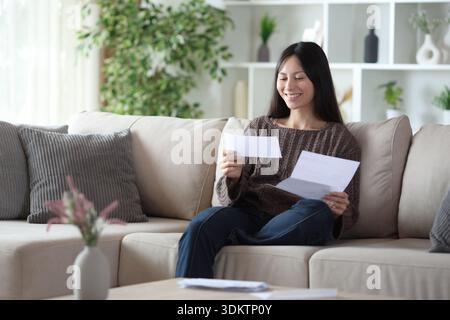 Happy asian woman reading letter sitting on a couch at home Stock Photo