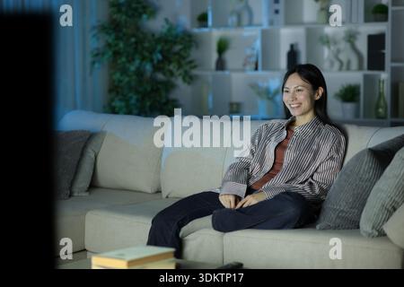 Single happy asian woman watching tv in the night sitting on a couch at home Stock Photo