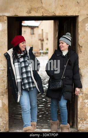Two women friends smiling and talking while exploring an old town in winter Stock Photo