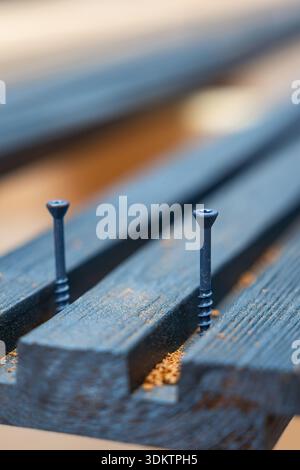 Two black screws partially embedded in dark wooden planks with sawdust scattered around, highlighting construction materials and craftsmanship. Stock Photo