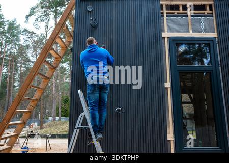 A worker in a blue hoodie adjusts components on a black paneled wall of a modern house with large windows, wooden framing, and a forested backdrop. Stock Photo