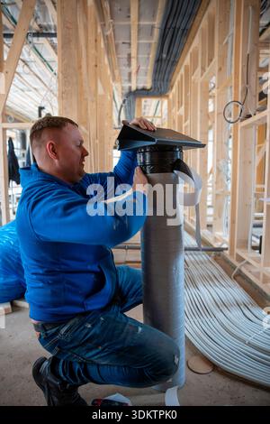 A worker in a blue hoodie secures insulated ductwork in a house under construction, with exposed wooden framing and coiled tubing on the floor. Stock Photo