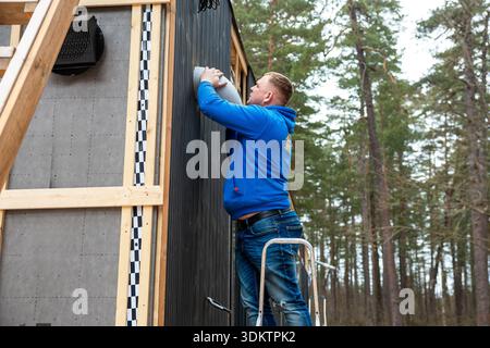 A man in a blue hoodie installs a ventilation system on the exterior wall of a partially constructed house with wooden framing, surrounded by pine tre Stock Photo