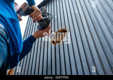 A worker in a blue hoodie uses a power drill on a black wooden wall, revealing insulation material. The scene highlights construction work on a new ho Stock Photo