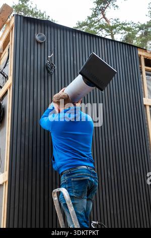 A worker in a blue hoodie installs a ventilation duct on a modern house with black paneling and wooden framing, surrounded by tall forest trees. Stock Photo