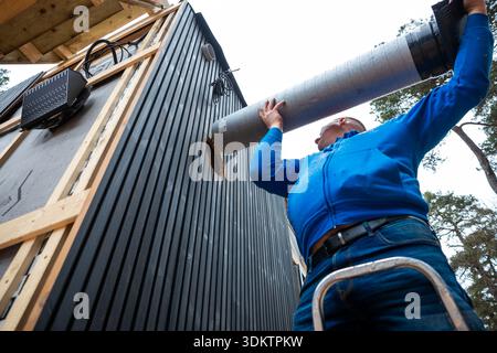 A worker in a blue jacket installs a large ventilation duct on a black paneled house under construction, framed with wooden beams near tall trees. Stock Photo