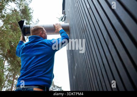 A worker in a blue sweatshirt installs a ventilation duct on a black wooden house with vertical paneling, surrounded by tall trees in a forested area. Stock Photo