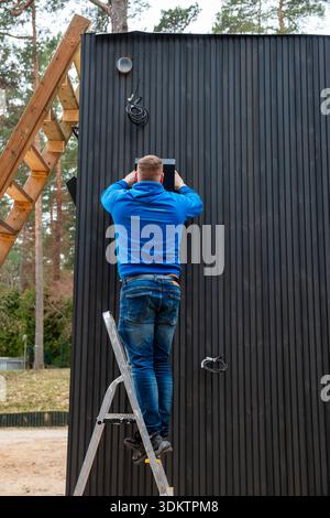 A man in a blue sweatshirt and jeans stands on a ladder adjusting a ventilation system on a black paneled building surrounded by tall trees. Stock Photo