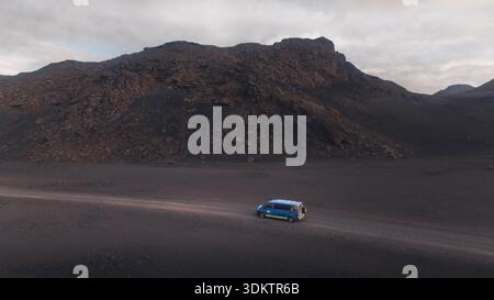 Aerial view of a blue van navigates the desolate, dark volcanic landscape near the mountains, contrasting starkly against the cloudy sky, Selfoss, Ice Stock Photo