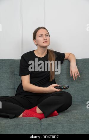 Bored young woman is sitting on a sofa with crossed legs, wearing red socks and a black t-shirt and pants, holding a remote control while watching tv. Stock Photo