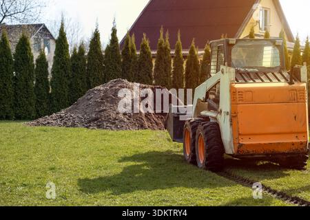 An orange and white skid steer loader sits on grass with tire tracks nearby. A soil pile and tall evergreen trees partially obscure a yellow house. Stock Photo
