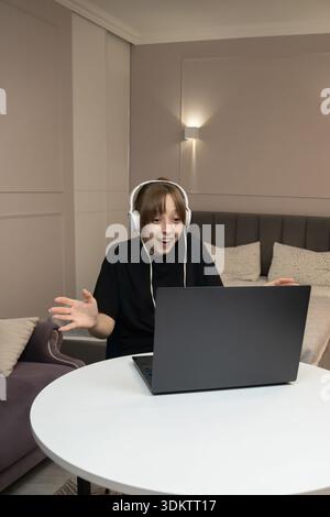 Enthusiastic woman in headphones spreading her arms to the sides while working on laptop, expressing joy and excitement during video call or online co Stock Photo