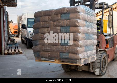 A forklift moves a pallet of plastic wrapped bags in an industrial area. A white van, orange cement mixer, and scattered wooden pallets are visible. Stock Photo