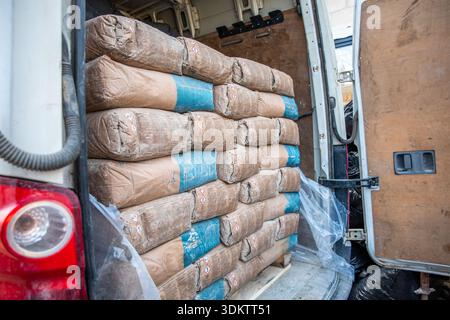 Open rear compartment of a white van with brown and blue cement bags stacked neatly. Wooden paneling and red taillight are visible, with plastic sheet Stock Photo