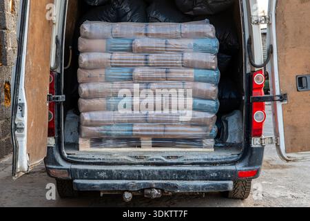 A stack of plastic wrapped cement bags on a wooden pallet inside a white delivery van. Black bags fill the rear space. Van parked on concrete near cin Stock Photo