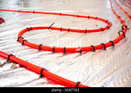 Bright red heating pipes are coiled and secured with black fasteners on a reflective foil insulation layer, highlighting the installation process. Stock Photo