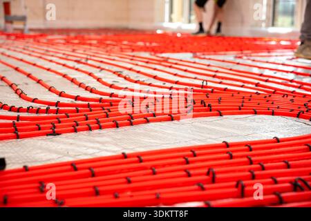 Bright red heating pipes arranged in a grid on reflective insulation, secured with black fasteners. Light walls, large windows, and workers' legs visi Stock Photo