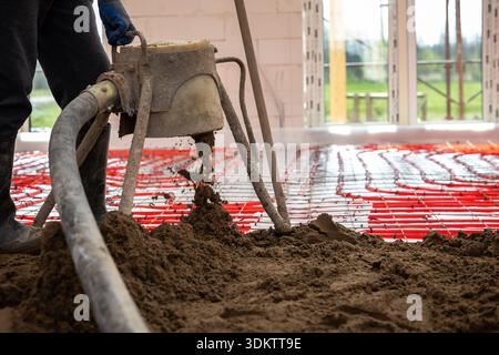 Red heating pipes are arranged on the floor as a worker pours screed concrete using a pump inside a building under construction with open windows. Stock Photo