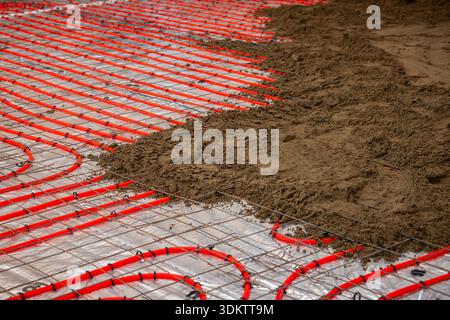 Red underfloor heating pipes are installed over reflective insulation, secured by a metal grid, with Estrich concrete partially covering the pipes. Stock Photo
