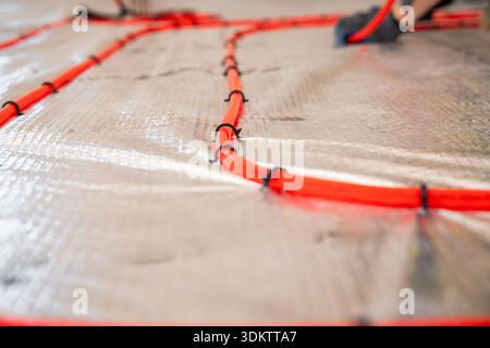 Red underfloor heating pipes secured with black clips on a reflective surface, with a worker's gloved hand visible during active installation. Stock Photo
