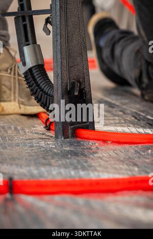 Red underfloor heating pipes are laid on a reflective surface, with a metal tool and a worker's shoe visible, capturing active installation work. Stock Photo
