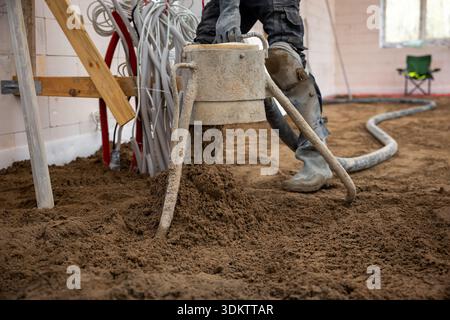 A construction worker pours and levels Estrich concrete over underfloor heating pipes in an unfinished indoor space with visible wiring and materials. Stock Photo