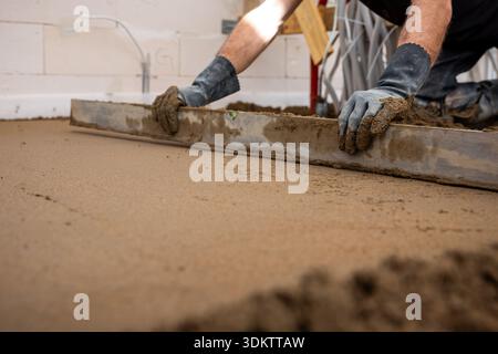 A worker in gloves uses a straightedge tool to level wet concrete indoors. White walls and construction materials are visible in the background. Stock Photo