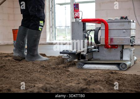 A construction worker operates a leveling machine with a red handle on concrete in an unfinished interior with white brick walls and large windows. Stock Photo