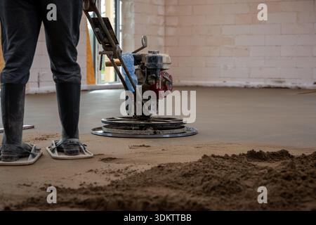 A worker operates a power trowel to smooth concrete flooring over underfloor heating pipes in a room with white brick walls and an open doorway. Stock Photo