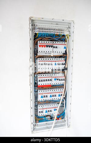 An open electrical panel embedded in a white wall, showing rows of circuit breakers, color coded wiring, and labeled switches in an organized layout. Stock Photo