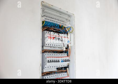 An open electrical panel embedded in a white wall, showing circuit breakers, blue, brown, and yellow green wires in an organized but unfinished state. Stock Photo