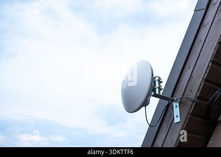 A white circular Wi Fi router is mounted on a wooden house exterior with visible cables and a metal bracket, set against a bright, partly cloudy sky. Stock Photo