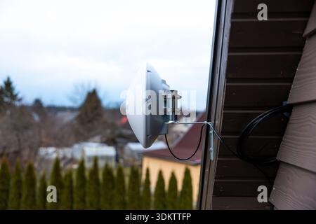 White dome shaped Wi Fi router mounted on house wall with metal brackets and cables, set against suburban homes, trees, and an overcast sky. Stock Photo