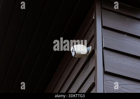 A white security camera is mounted on a dark brown wooden wall with horizontal panels at a private house, positioned to monitor the surroundings. Stock Photo