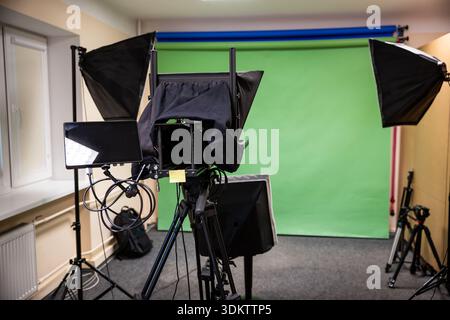 A professional studio setup with a green screen, camera on tripod, softbox lights, monitor, cables, beige walls, window, radiator, and carpeted floor. Stock Photo