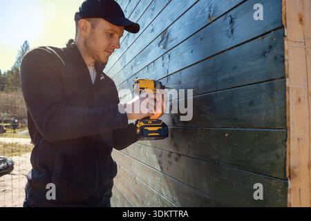 A man in a black cap and jacket works with a yellow and black power drill on a polished wooden wall. Blurred trees and open space are visible in sunli Stock Photo