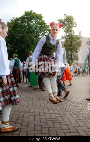 Children in traditional Latvian costumes gather outdoors on cobblestone ground. A girl mid jump wears a plaid skirt, white blouse, and green vest. Stock Photo