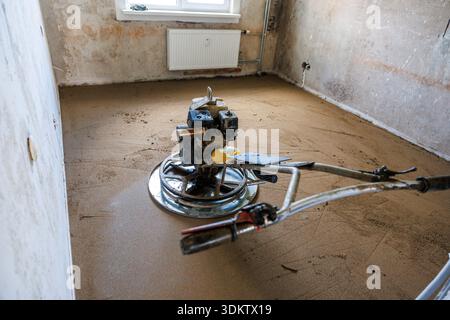 A construction site shows a room with unfinished walls, a floor screeding machine on fresh concrete, a radiator below a window, and visible wall pipes Stock Photo