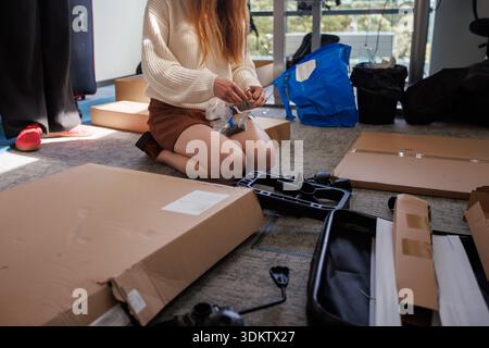 Individual in white sweater and brown skirt kneels on carpet, assembling items from boxes. Black case, hardware pieces, and blue bag are visible nearb Stock Photo