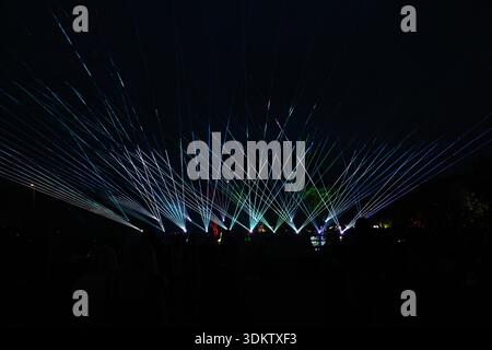 Laser beams in blue, green, and white radiate outward in a fan like pattern against a dark sky, with silhouettes of people and faint trees visible. Stock Photo