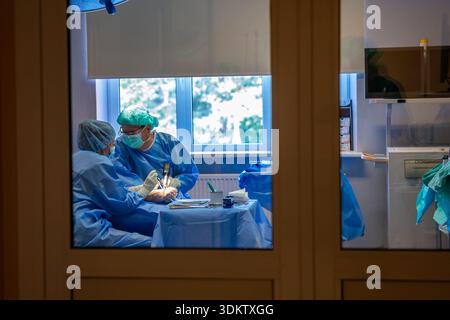 Two professionals in blue surgical attire work in a well lit operating room with visible equipment, cabinets, and a monitor, viewed through a glass do Stock Photo