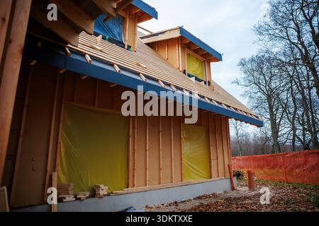 Builders work on a timber house in a fenced site with leafless trees. Exposed studs, two dormers, blue eave flashing, yellow sealed openings, and roof Stock Photo