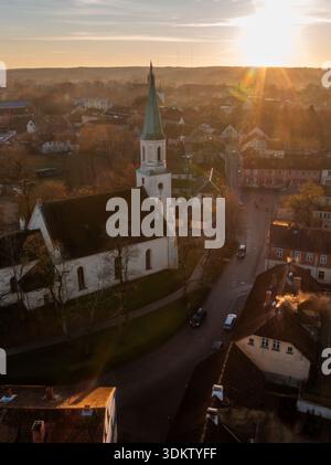 St. Catherine's Church catches warm light as narrow streets curve by red roofs in Kuldiga, Latvia. Chimney smoke rises, long shadows stretch, soft haz Stock Photo