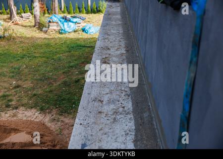 A construction site shows a concrete ledge on a gray building, with grass, sand, blue tarps, wooden planks, trees, and a fence in the distance. Stock Photo