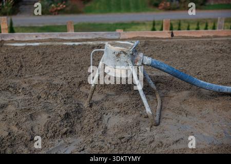 A concrete leveling tool with a blue hose rests on wet concrete, surrounded by wooden planks. A paved pathway with grass and plants is visible nearby. Stock Photo