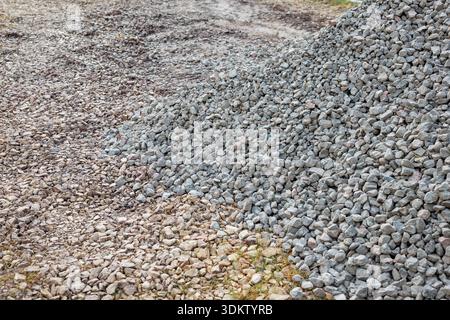 A large pile of small gray stones rests on uneven dirt and gravel, surrounded by scattered rocks and patches of grass, suggesting a construction site. Stock Photo