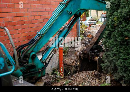 A blue excavator digs near a red brick wall with an orange pipe exposed. Gravel, tools, and a tree partially obscuring the bucket are visible. Stock Photo