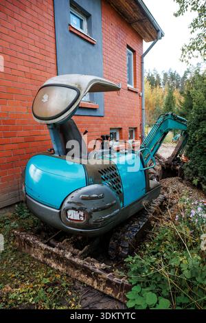 A worn blue excavator is parked by a red brick building with a wooden roof overhang, near a gravel filled trench, surrounded by plants and bushes. Stock Photo